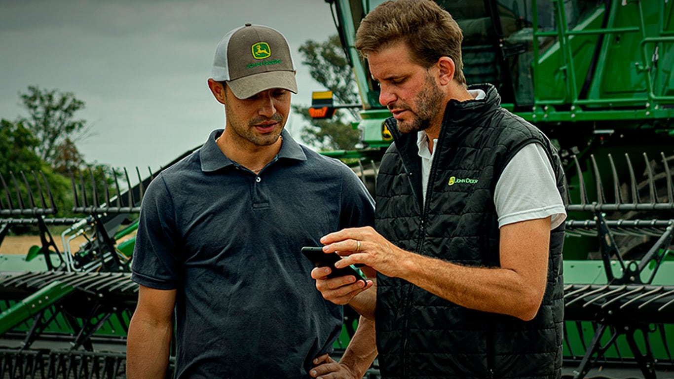 Dos hombres manipulando un teléfono celular en el campo frente a una cosechadora John Deere