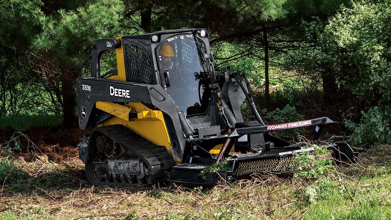 Segadora Giratoria en un Cargador Compacto sobre Orugas 333E trabajando cerca de un bosque.