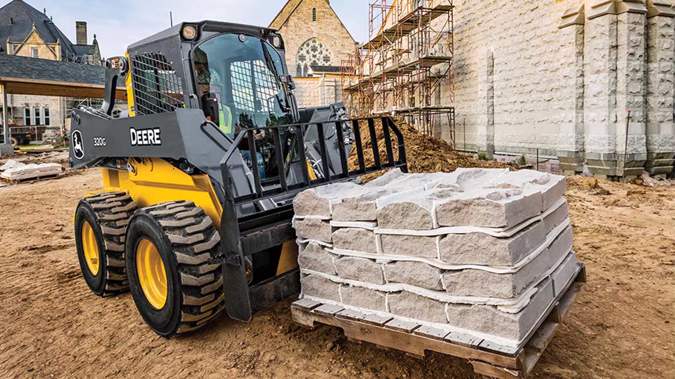 El minicargador John Deere 320G lleva bloques de piedra en un sitio de construcción con andamios.