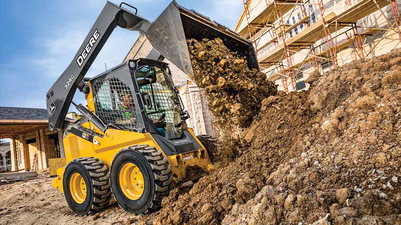 El minicargador deslizante John Deere amarillo descarga tierra en un sitio de construcción con andamios.