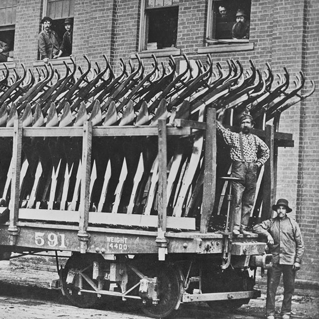 Fotografía de 1882 de tres hombres parados junto al vagón del tren de Deere & Co. cargado con arados de acero listos para ser despachados; se ven también unos empleados de la fábrica mirando por la ventana del edificio de atrás