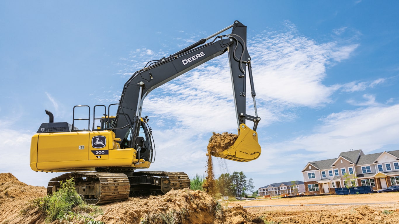 Una Excavadora 200G mueve tierra en un lugar de trabajo con un desarrollo habitacional en el fondo.