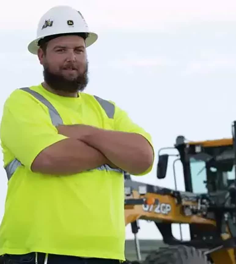 Ryan de pie frente a la maquinaria de construcción Deere con los brazos cruzados