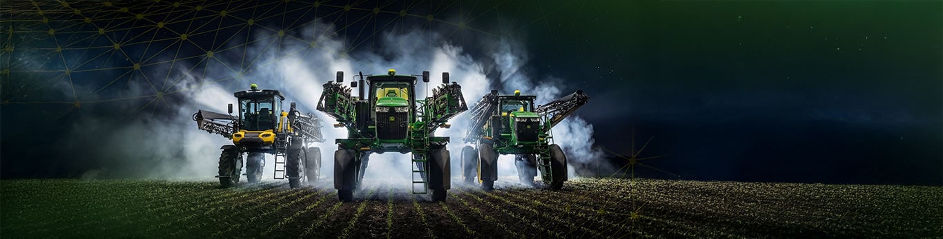 2 pulverizadores John Deere en el campo con humo en el fondo.