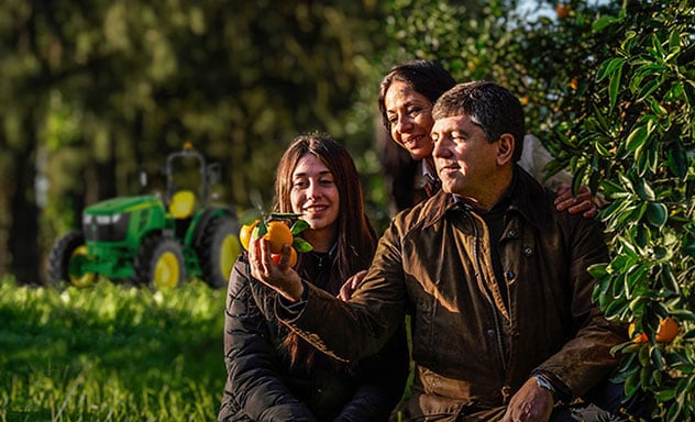 Familia unida y sonriendo en el campo, con un tractor John Deere al fondo de la imagen.