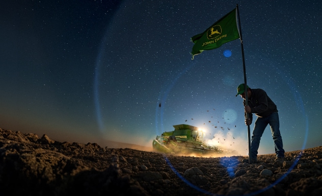Un hombre colocando una bandera de John Deere en el suelo, una cosechadora al fondo.