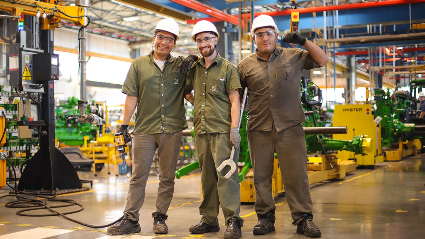 Tres hombres uno al lado del otro, usando equipos de protección individual dentro de un taller de máquinas John Deere.