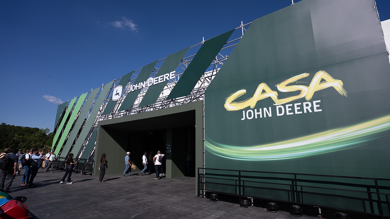 Entrada de carpa John Deere verde en evento al aire libre, con paneles diagonales y personas entrando bajo cielo azul.