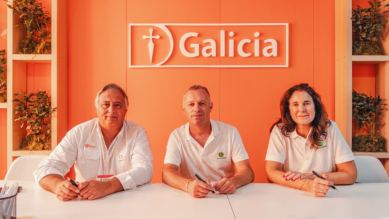 3 personas, 2 hombres y una mujer, sentados en una mesa frente a una pared naranja con el logo Galicia.