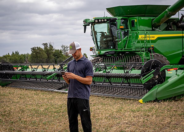 Un hombre en el campo, manipulando su teléfono celular. Al fondo, una cosechadora S7.