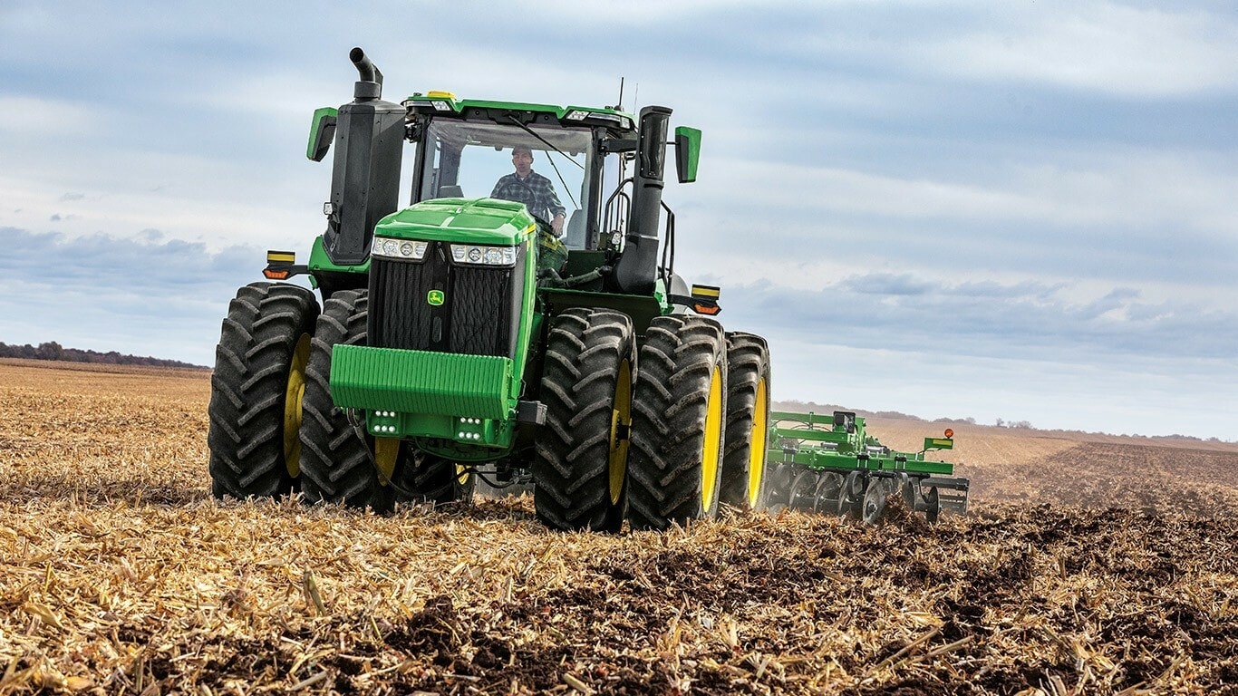 Un tractor agrícola John Deere de gran tamaño con neumáticos dobles, operando en un campo arado bajo un cielo nublado.